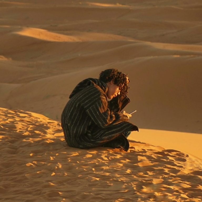 Eine Person in traditioneller Kleidung sitzt nachdenklich auf sandigem Terrain.