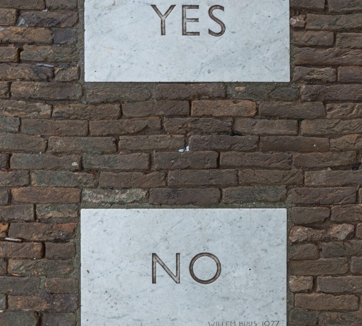 Graue Steintafeln mit der Aufschrift "YES" oben und "NO" darunter, vor einer Mauer.