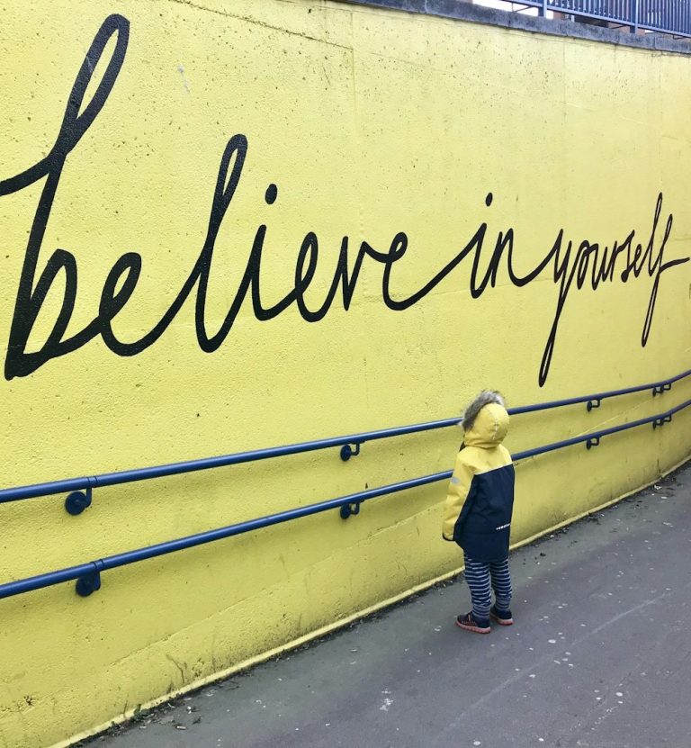 Ein Kind in gelbem Regenmantel steht vor einer gelben Wand mit der Aufschrift „Believe in yourself“.